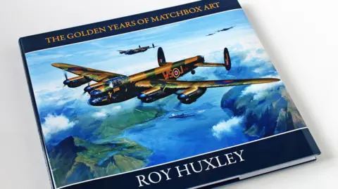 The cover of a hard-backed book. The cover features a painting of four military aircraft flying over blue water, white blobs of cloud, and green hills. A ship is sailing on the water below the planes. Across the top of the cover are the words: "THE GOLDEN YEARS OF MATCHBOX ART", written in yellow on a blue background. Across the bottom are the words: "ROY HUXLEY", written in white on a blue background.