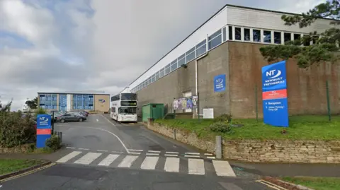 The front of Newquay Tretherras School, with two blue signs with the school name in white, and two brick buildings on the perimeter of a car park, in which a double decker bus is parked