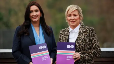 PA Media Emma Little-Pengelly, left, is holding a copy of the strategy. She is smiling at the camera and has long brown hair. She is wearing a navy suit jacket with a light blue silk scarf. Michelle O'Neill, right, is also holding the strategy. She is wearing a leopard print blazer and white shirt. Her hair is blonde and tied back.