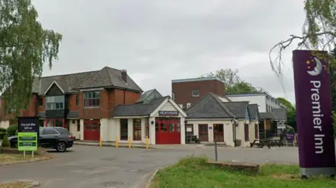 A Premier Inn hotel made up of a series of buildings. They are partially painted white and partially red brick. There is a car park in front and purple and green signage.