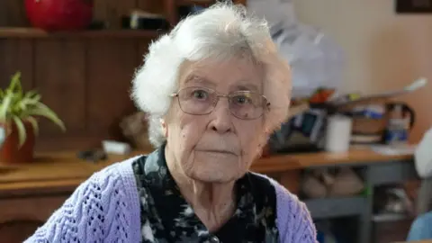 Shaun Whitmore/BBC Heather Manthorpe is sitting down and looking directly at the camera. She is wearing a black patterned blouse, a purple knitted cardigan and glasses.