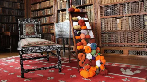National Trust/Camille Francois Dozens of colourful knitted pumpkins covering a wooden ladder in an old library, which stands next to metal chair. Hundreds of books line the walls of the rooms, with red carpet on the floor.