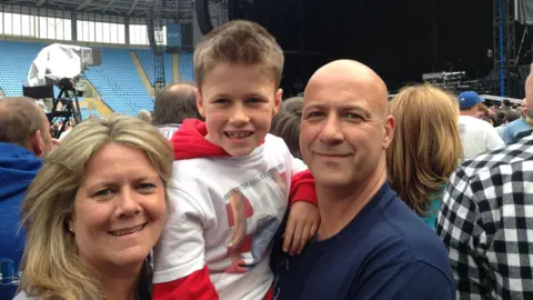 Family photo A young boy smiles at the camera. He has a red hoody on and a white t-shirt over the top. He is being held up by a man who has a blue t-shirt and a bald head who is also looking at the camera. On the other side of him is a woman with blonde hair and a grey t-shirt. They are all cuddled up posing for a photo, behind them are fans in a stadium all facing towards the stage, which can be seen at the back of the photo.