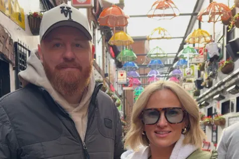 A man and woman are looking into the camera. the man on the left is wearing a white baseball cap, a white hoody and a grey zip up jacket. He has a ginger beard. The woman has shoulder-length blond hair, a grey hoody, green coat and and tortoiseshell glasses. Behind them is a number of colours umbrellas hanging from a metal grid.