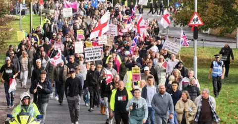 A number of people at a protest in Crowborough, East Sussex, on 8 November. Demonstrators are resisting plans to house asylum seekers at the former army camp in the town. A number of England and union jack flags can be seen in the image, alongside a few placards.