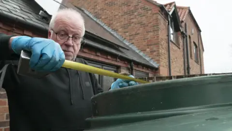 Bob Hayes, who has grey hair and black-rimmed glasses, uses a tape measure to check the oil level in his tank.