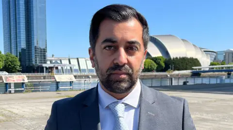 Humza Yousaf in a grey suit and white shirt in front of the Quayside in Glasgow.