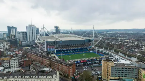 Aerial shot of the Principality stadium in Cardiff. The river taff runs through the city on the right hand side, while the green Cardiff Arms Park football pitch is in front of the stadium. High rise buildings and houses surround the stadium. 
