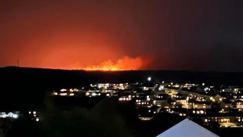 Gregg Portass Flames from a fire on Rosenannon Downs seen from Wadebridge during the night. Dozens of houses can be seen in the foreground with their lights on. Flames and smokes are going up into the night sky with an orange glow around the site.