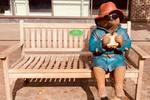 TAG A Paddington Bear sculpture eating a marmalade sandwich on a bench. Someone has placed sunglasses on him.