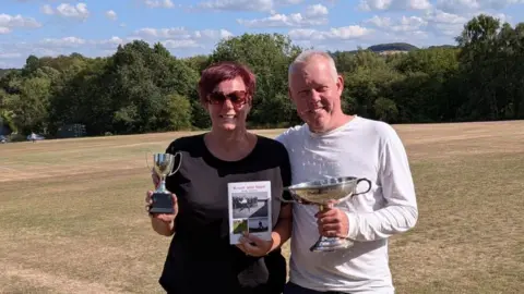 Boz Davison A woman with short red hair and a man with short white hair stands beside one another on a field. They are holding trophies