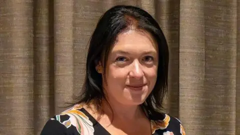 Wiltshire Search & Rescue Head and shoulders image of Joanne Young with shoulder length black hair and wearing a black, floral top. She is standing in front of a brown curtain.