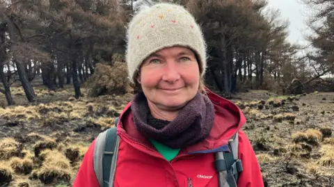 Carla Fowler/BBC A woman with green eyes wearing a grey woolly hat with different coloured hearts on it. She is wearing a burgundy scarf and a red waterproof coat. Behind her is brown moorland and a small forest.