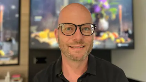 Nick Baynes looks to the camera, he is wearing a dark shirt and glasses. Behind him are colourful screens showing the game's menu