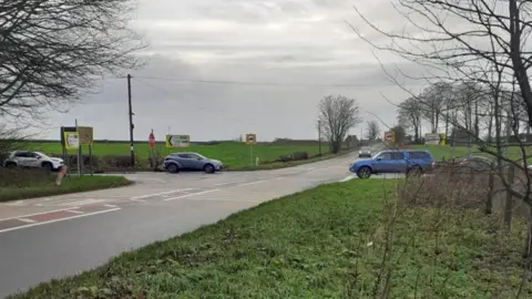 A four-way junction in the Somerset countryside surrounded by fields. Cars are waiting to pull onto the bigger road. 