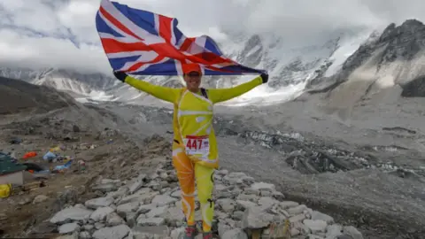 Anuj Adhikary A Sally Orange wearing a yellow and orange citrus costume while hold the union Jack flag on Mount Everest.