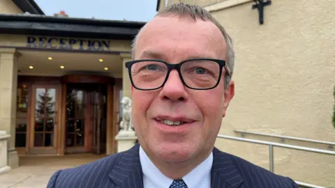 Tim Boyd has black square glasses and is wearing a striped blue suit. He has a light blue shirt and grey hair. He is clean shaved. Behind him is a yellow building and entrance.