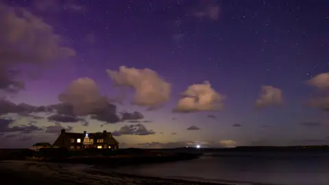 Jonathan A house sits on the beach, the sky above is a purple glow, not the traditional appearance of the northern lights