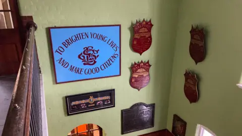 A collection of plaques and signs hung against a light-green painted wall within Salford Lad's Club. One reads 'To brighten young lives and make good citizens', which is the club's motto. 