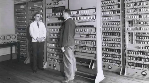 Cavendish Laboratory A black and white image shows two men standing in front of a bank of old computers.