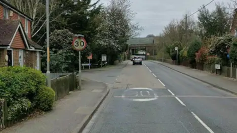 A road with cars and trees on it, a 30 MPH sign and a bridge in the distance. 
