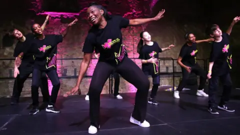 James O'Jenkins A group of young dancers, wearing black yellow and pink tops with black trousers, perform on stage