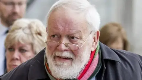 Michael Longley staring into the left hand side of the picture. He has white hair and a white beard with gold glasses perched on his chin. He is wearing a dark coat and a red, pink and red striped scarf