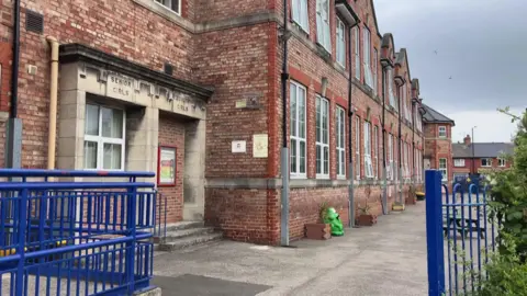 A red-brick building with large white windows and a stone doorway to the left. An open, blue gate and blue fencing marks the entrance to the school, with a large playground to the right immediately in front of the school building. It is a cloudy day.