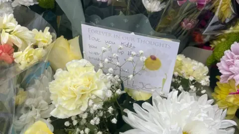 A handwritten message is tucked into a bouquet of flowers saying "love you kiddo, chill up there with your nan"