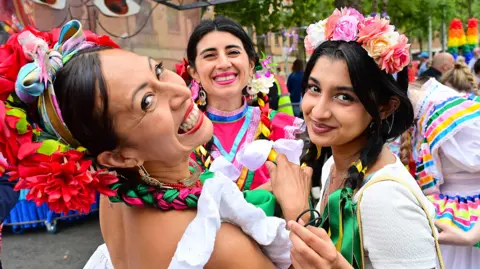 Three woman are smiling at the camera. They have black hair and are wearing colourful clothes. 