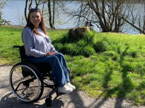 Eva Hanna Eva Hanna sits in her wheelchair in front of grass and some water with bare trees in the background and the sun shining
