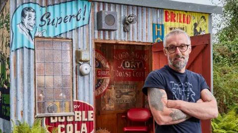 A man in a navy blue T-shirt and jeans stands in front of a corrugated shed with colourful painted slogans