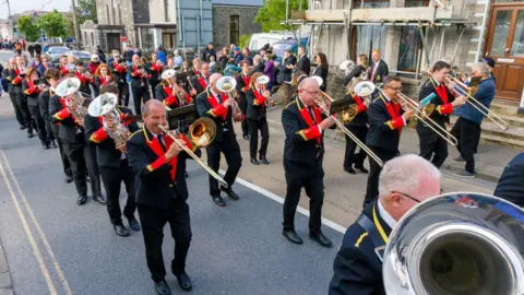 St Dennis Band A brass band marches along a road while playing their instruments. People are stood on the pavement watching.