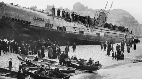 Getty Images A black and white archive photo of a submarine in shallow water on Hastings Rocky Beach. People are on top of the vessel and others are on the beach watching the craft. Some are in boats on the shore.