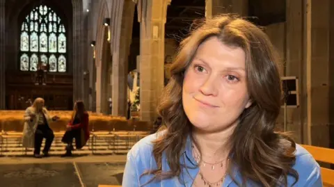 Amy Van Zyl has shoulder length, brown wavy hair and is wearing a blue shirt with a silver necklace. The main central area of St Nicholas Cathedral is behind her with two women sitting on seats and talking in the background.