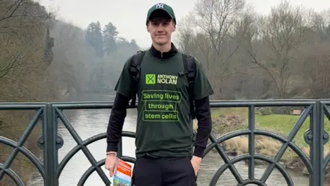 Family A young man standing on a bridge over a river. He is wearing a green New York Yankees baseball cap and a green T-shirt which reads: "ANTHONY NOLAN - Saving lives through stem cells". He has a rucksack on his back and is holding what looks like a map.