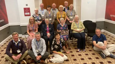 Piers Hopkirk/BBC 14 former pupils from Loose Primary School from 1966 plus their teacher (in the centre holding photo) Shirley Arnold pose in four rows in a room  