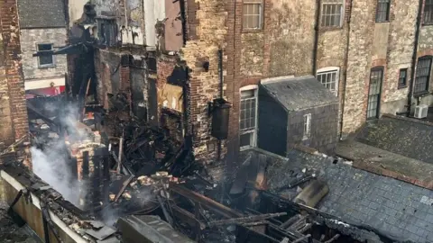 Smoking timber and pies of charred rubble spread along the ground from a building in the centre of a brick terrace.