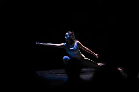 a contemporary dancer onstage, reaching with her hand turned towards the ceiling; one of her legs is in a deep squat (stock image)