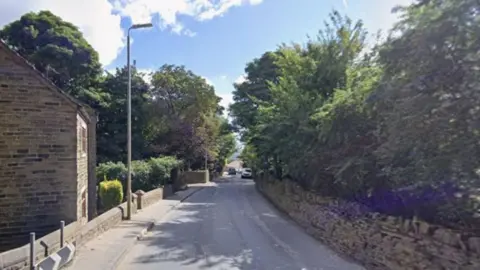 Google A quiet, slightly winding road bordered by stone walls and dense greenery on both sides. On the left, there is a building made of traditional stone with a sloped roof. A narrow pavement runs alongside it, separated from the road by short metal posts. Trees line both sides of the road. A tall streetlamp stands near the centre-left of the scene. In the distance, the road slopes gently upward toward a small cluster of buildings and a parked vehicle, partially visible between the trees.