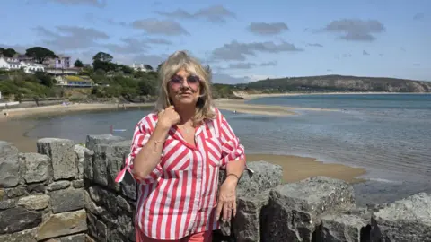 A blonde woman in a red and white striped shirt stands in front of a wall overlooking the seaside 