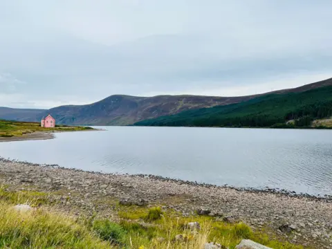 Roseanna Mitchell Clear sky above a still loch, surrounded by hills and a bright pink building visible in the distance