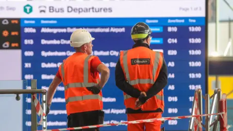 Pacemaker Two men with backs to the camera. They are wearing hi-vis jackets and hard hats, standing in front of a departures board showing bus routes and a timetable.