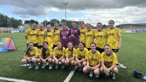 Finn Harps team standing for a photo in their yellow jerseys, the goalkeepers are wearing burgundy jerseys. All the girls have their hair tied back and are smiling at the camera whilst on the football pitch.