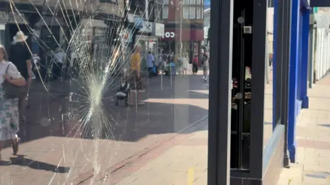 A view of a shop front window that has been smashed. People can be seen walking past in the window reflection. 