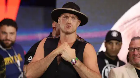 Tyson Fury in a black hat and vest holds up his fists
