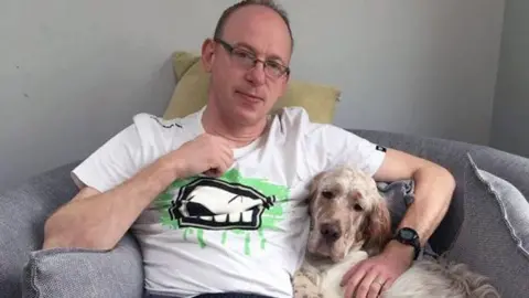 A man in glasses and a white t-shirt sits holding a dog

