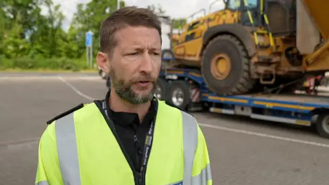 BBC/Gary Darfield An officer in a high-vis yellow jacket and black fleece stands in a car park in front of an HGV trailer carrying heavy machinery. He has short brown hair and a greying beard. His mouth is slightly open and he appears to be in the middle of speaking.