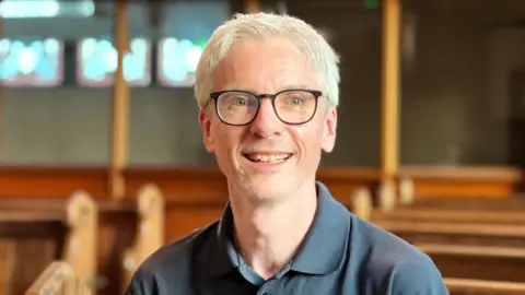 A man with white hair and black thick-rimmed glasses smiles at the camera as he sits inside a church.