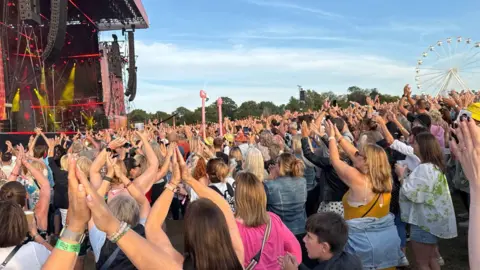 Lauren Carter/BBC A packed crowd of people clapping along to a song. There is a big stage to the left and a Ferris wheel in the background.